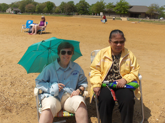 Dona and Nanta are sitting in the sun on beach chairs and smiling.  Dona is holding an umbrella to give her shade and is wearing short pants and a long-sleeved shirt, Nanta is wearing a jacket and long pants.
